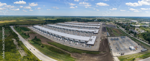 Aerial panorama of a large industrial data center complex with long buildings, loading docks, and an electrical substation near green fields