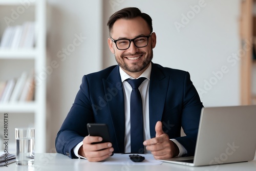 Confident businessman wearing glasses and suit holding smartphone at office desk smiling at camera