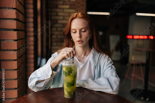 Bild auf Leinwand Portrait of redhead young woman sitting at cafe table, stirring refreshing mojito like drink with straw, enjoying moment of leisure and relaxation during summer
