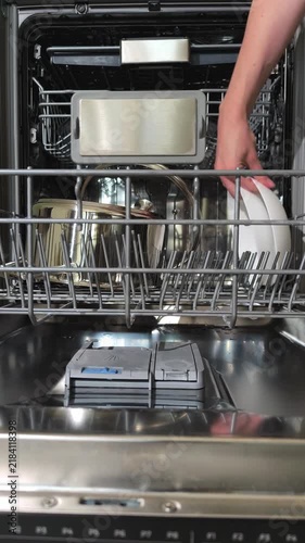 Close up hands removing clean plates from dishwasher lower rack. Person unloads washed dishware from open basket in tidy home kitchen. Vertical video captures dish organizing during cleaning