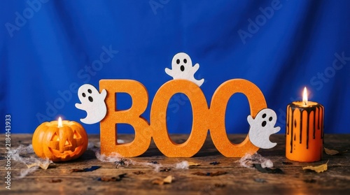 Halloween decoration with orange boo sign, ghosts, pumpkin, and dripping candle on a rustic wooden table against a blue background