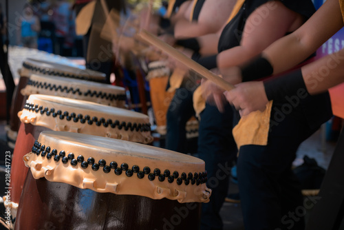 A group of taiko drummers performs outdoors, striking traditional Japanese drums with wooden bachi sticks in a coordinated, energetic ensemble performance.