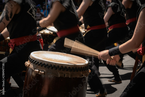 A group of taiko drummers performs outdoors, striking traditional Japanese drums with wooden bachi sticks in a coordinated, energetic ensemble performance.