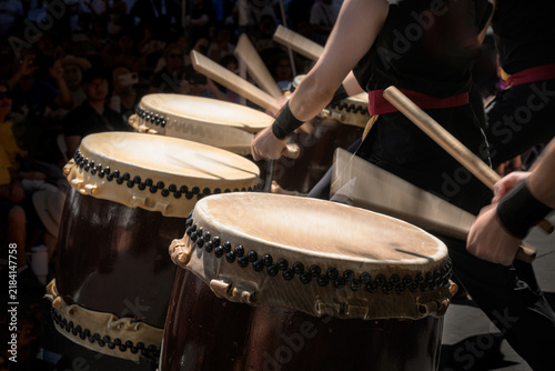 A group of taiko drummers performs outdoors, striking traditional Japanese drums with wooden bachi sticks in a coordinated, energetic ensemble performance.
