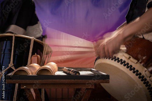 A group of taiko drummers performs outdoors, striking traditional Japanese drums with wooden bachi sticks in a coordinated, energetic ensemble performance.