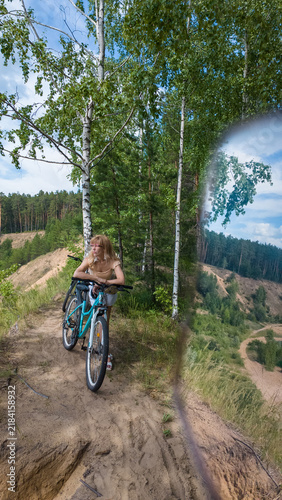 A girl is riding her bicycle in a forest path surrounded by tall trees. The sun shines brightly through the leaves, creating reflections in the scene.