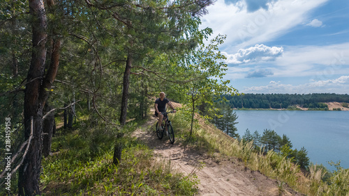 A man is biking on a dirt path near a lake surrounded by trees. The sun is shining, and the sky is clear with some clouds. The scene shows a natural setting with water and greenery.