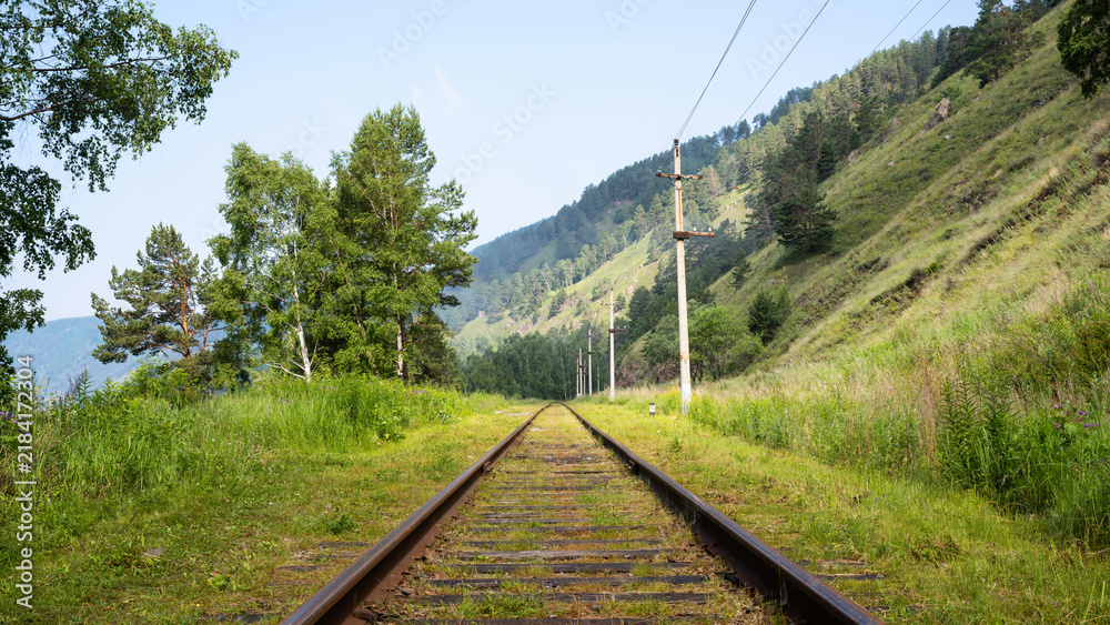 Obraz premium Empty railway track stretching into the distance through a green summer landscape, Lake Baikal, Russia. Historical rural train line with mountains and trees. Journey and travel concept