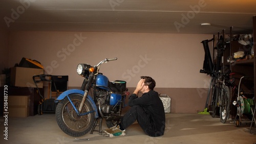 Canvas Print Young man sitting on the garage floor, covering his face with hands, experiencin