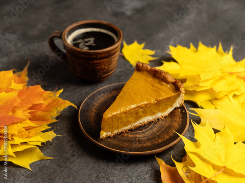 A slice of pumpkin pie on the table with a cup of coffee. Autumn yellow maple leaves. A traditional dessert on Thanksgiving.