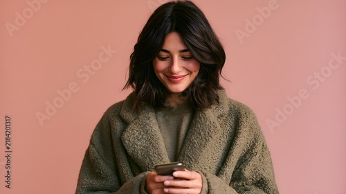 Smiling young woman with dark hair wearing green coat looks down while holding smartphone against plain solid pink studio background