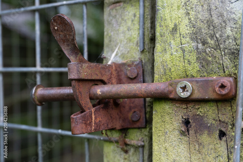 Close-up of a heavily rusted iron auto-latch and sliding bolt mechanism securing a wire mesh farm gate. The latch is mounted to a weathered, green moss-covered wooden fence post with screws.
