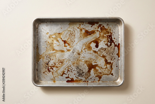 Top view of a dirty greasy baking sheet on a beige background. Unwashed aluminum metal pan covered in burnt food residue and oil. Flat lay of messy kitchenware