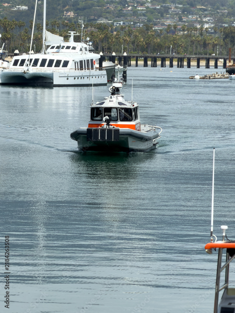 Fototapeta premium SANTA BARBARA, CA, APR 9, 2026: Harbor Patrol response boat motoring through the harbor channel, leaving a wake, with a catamaran yacht and pier behind