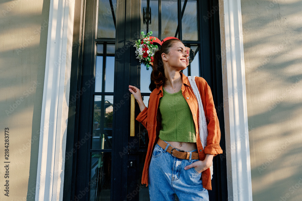 Obraz premium Smiling young woman in an orange shirt and jeans stands by a black door with a festive wreath, holding a bag. Concept of holidays and winter mood.