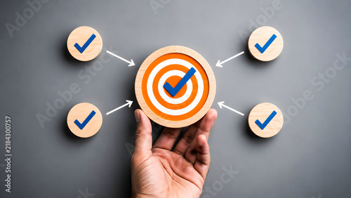 Hand holding wooden block with orange target and blue checkmark symbol surrounded by four smaller circular wooden icons with checkmarks pointing towards center on dark grey background for business str
