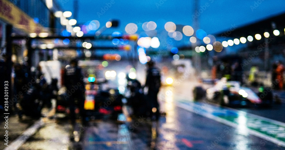 Obraz premium A blurred image of a racetrack during a nighttime Formula 1 event, featuring pit crews and cars under lights amidst rainy conditions.