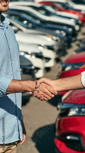 Car Dealership Handshake: A close-up shot captures two men shaking hands amidst a row of new cars at a dealership, symbolizing a successful transaction and a new beginning.