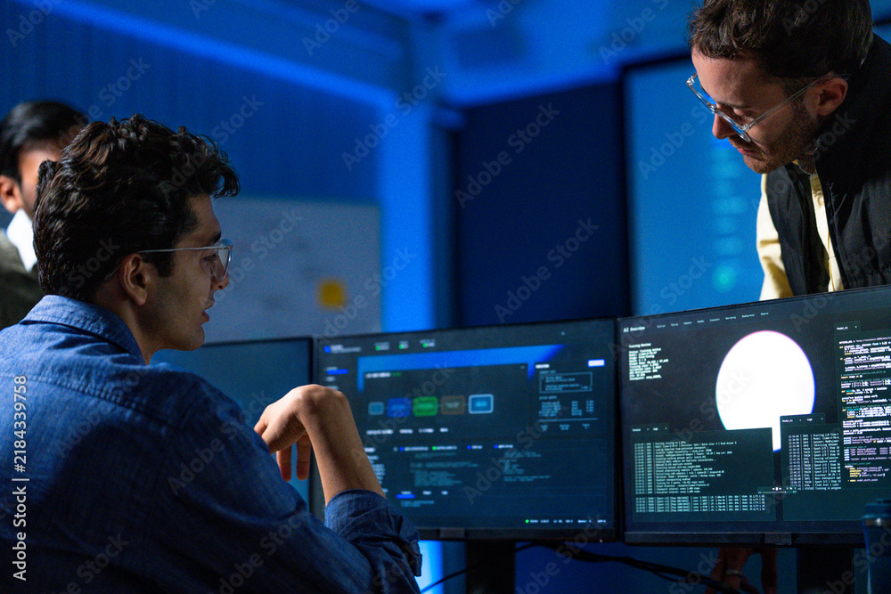 Fototapeta premium Two male colleagues of different generations working through a problem points at a line on the monitor while the younger engineer listens in a night shift operations room