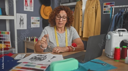 Woman dressmaker sketching garment designs on paper, right hand drawing near laptop with tape measure and pincushion in studio; focused creativity.