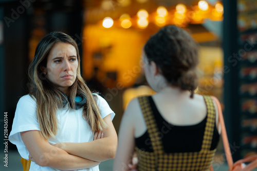 Disappointed Sad Woman Listening To Friend During Serious Outdoor Conversation. Unhappy person stands with folded arms while interacting with another girl