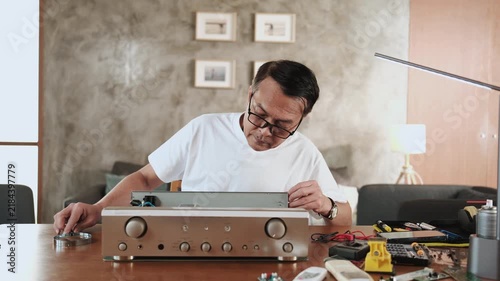 Mature Asian man disassembling and checking audio equipment, looking with magnifying glass at circuit board details for repair, home appliance DIY fix, electronics component hobby, handyman workshop.