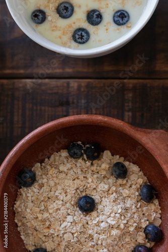 Aerial view of a rustic clay bowl filled with oats and blueberries next to a cup of yogurt with oats and blueberries on a rustic table