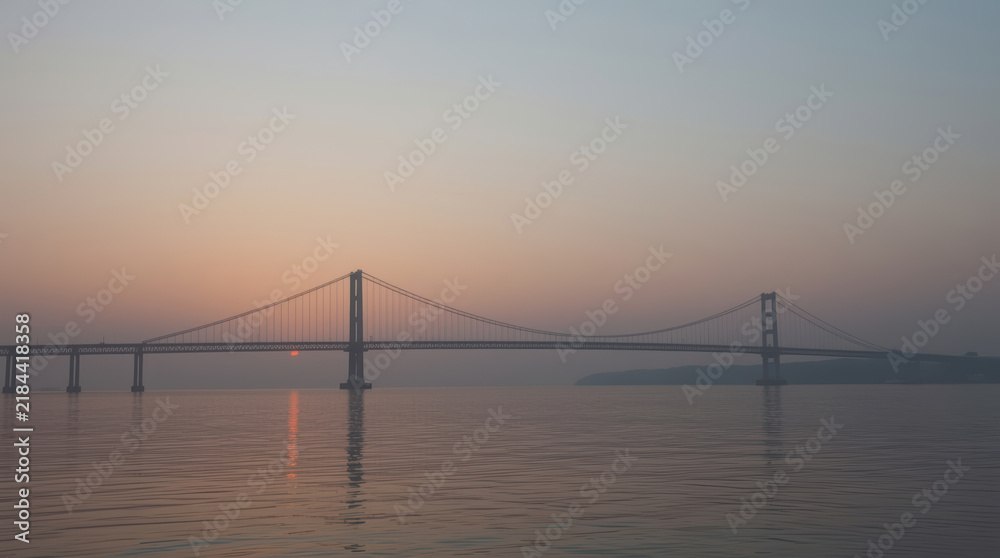 Obraz premium Suspension bridge over calm water beneath hazy sunset sky