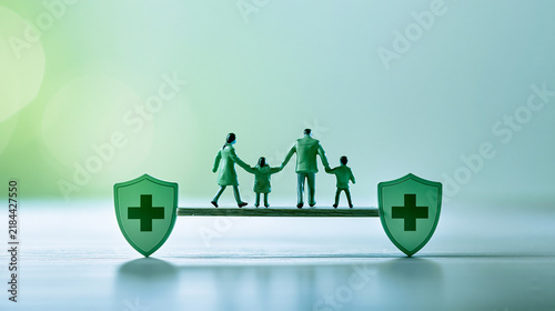 A miniature family of four holding hands stands on a balance beam supported by two shields with medical crosses, symbolizing health, protection, and family safety.