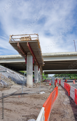Bridge construction site on a highway overpass featuring scaffolding, concrete pillars, orange traffic barriers and cones, urban infrastructure work and road maintenance under a blue sky with clouds.
