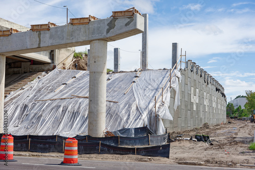 Bridge construction site on a highway overpass featuring scaffolding, concrete pillars, orange traffic barriers and cones, urban infrastructure work and road maintenance under a blue sky with clouds.
