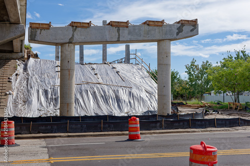 Bridge construction site on a highway overpass featuring scaffolding, concrete pillars, orange traffic barriers and cones, urban infrastructure work and road maintenance under a blue sky with clouds.
