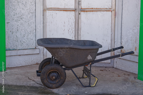 An empty, weathered wheelbarrow with rusty tires and metal frame parked on cracked concrete beside a peeling, white shed; evokes outdoor work, gardening, construction and vintage utility equipment.
