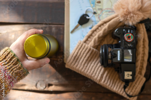 Rear view of a hand holding a metal water bottle on a rustic wooden table next to travel items, a map, a camera, and a wool hat