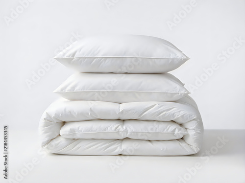 A stack of two fluffy white pillows and a folded white down comforter isolated on white background