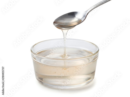 A clear, viscous liquid syrup pouring from a metal spoon into a small glass bowl, isolated on white background
