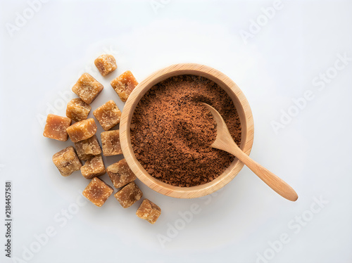 Top view of jaggery cubes and a wooden bowl filled with powdered jaggery with a wooden spoon isolated on white background