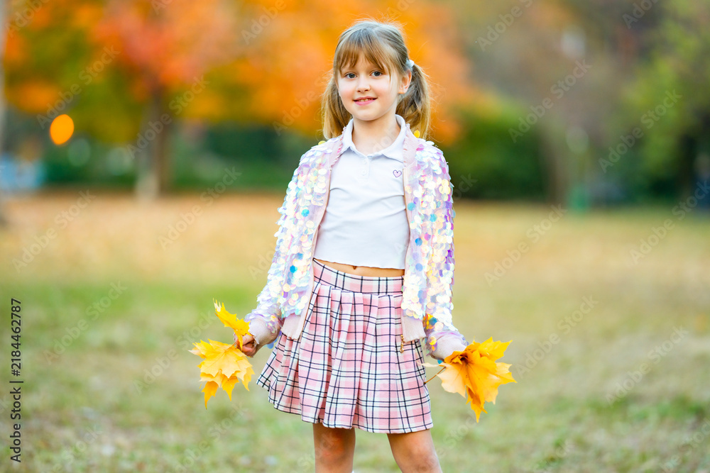 Fototapeta premium Child girl holds yellow leaves. Child wears sparkling jacket. Smiling child girl stands in park. Cute child portrait outdoors.