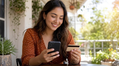 Smiling Hispanic woman tapping on a plain black smartphone while holding a credit card on a sunlit plant-decorated terrace during a warm afternoon lifestyle moment
