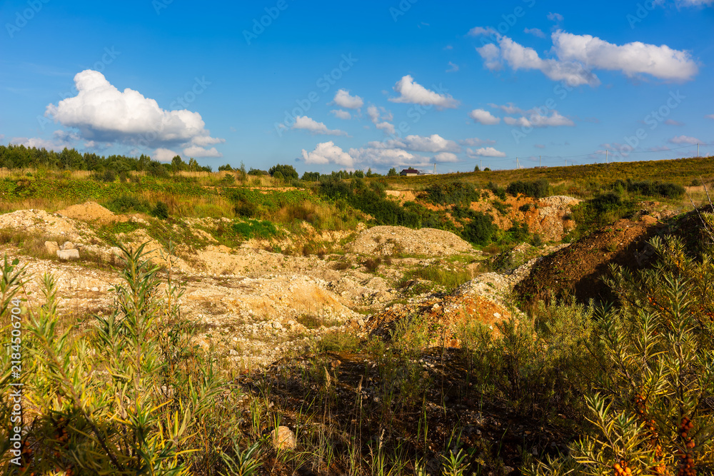 Obraz premium Rugged terrain of an abandoned sandy quarry with dry vegetation and loose rocks under a bright blue summer sky with fluffy white clouds on a sunny day in the countryside