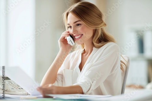 Smiling woman in a white blouse talking on the phone while reviewing documents in an office