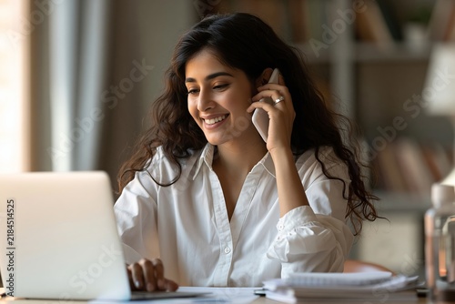 Young woman working on her laptop while talking on a mobile phone in a bright home office