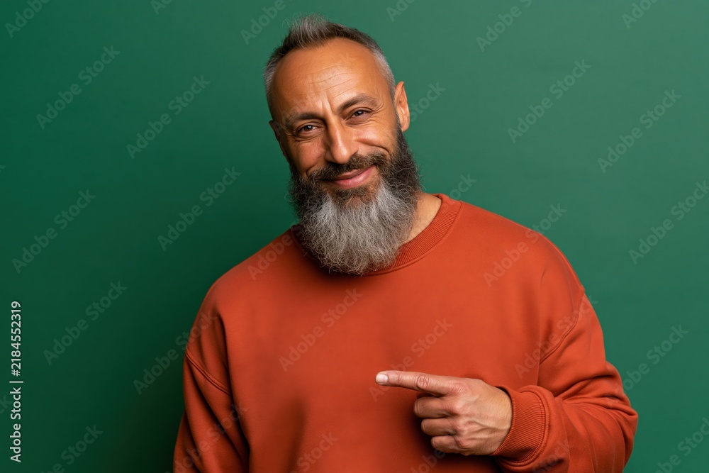 Fototapeta premium Gray bearded man pointing to the side in orange sweatshirt studio portrait on green background for lifestyle headshot and product endorsement