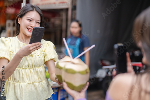 Asian woman using smartphone recording mobile video of fresh coconuts for street food review, Food vlogger, social media content creator, travel influencer and authentic local thai market tourism