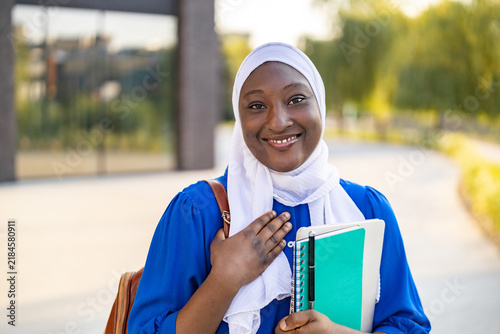 A happy African American Muslim woman wearing a hijab smiles while holding books and a tablet outdoors