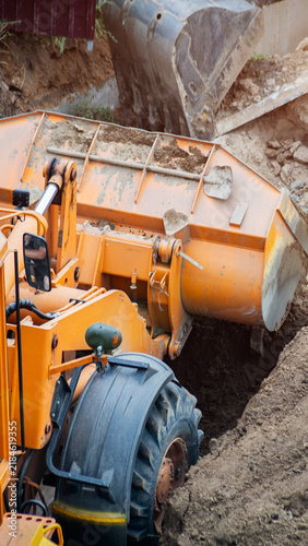 Orange construction loader bucket full of soil engaged in earthwork at construction site during day, heavy machinery in action moving ground for foundation work, vertical photo