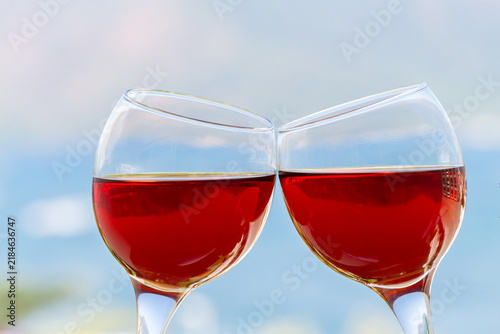 Close view of two stemmed glasses of rose wine leaning together in a toast with a softly blurred blue background suggesting a relaxed outdoor setting