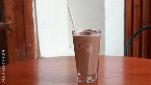 Chocolate milkshake sits on a wooden table. It holds a white straw and is in a tall glass. Behind it, a vintage wooden door with arched panels. The background shows weathered green and yellow paint
