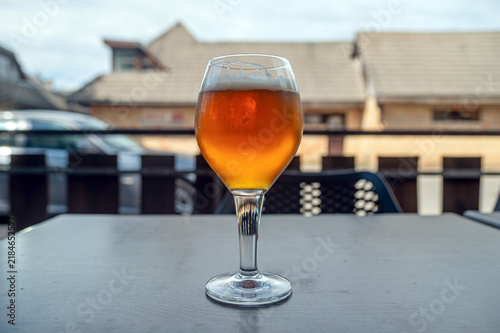 A glass of beer sits on a table outside a cafe during the day while cars pass by.