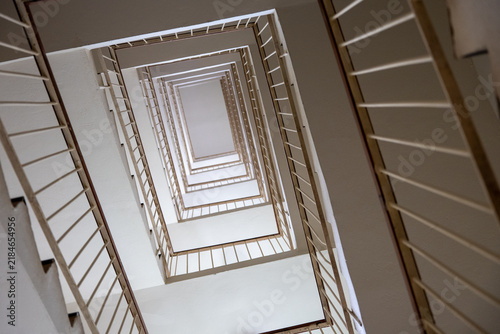 Quadro su tela Upward view through a multistory square stairwell with repeating metal railings, creating strong geometric perspective, symmetry, depth, leading lines and abstract architecture